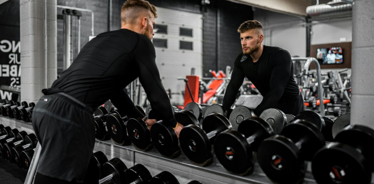 Man looking in mirror while standing over dumbbells in gym, representing strength training and focus