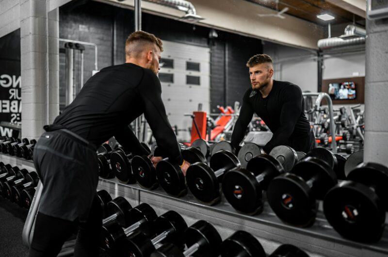 Man looking in mirror while standing over dumbbells in gym, representing strength training and focus